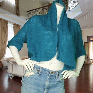 May include: Teal-colored, open-front knit bolero sweater with short sleeves. The sweater is displayed on a mannequin, paired with light-wash denim jeans. The bolero has a wide collar and a loose, airy knit.