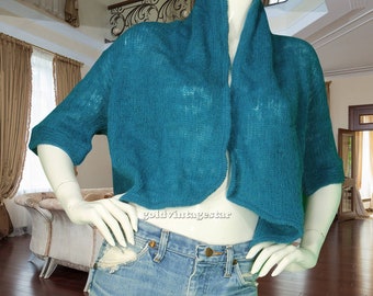 May include: Teal-colored, open-front knit bolero sweater with short sleeves. The sweater is displayed on a mannequin, paired with light-wash denim jeans. The bolero has a wide collar and a loose, airy knit.