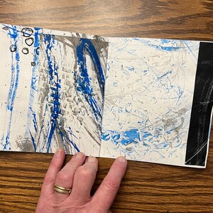 May include: A handmade book with blue and gray abstract art on the cover. The book is open to reveal more abstract art on the inside pages.