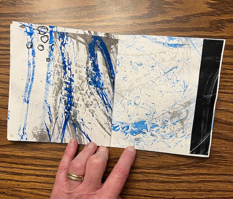 May include: A handmade book with blue and gray abstract art on the cover. The book is open to reveal more abstract art on the inside pages.