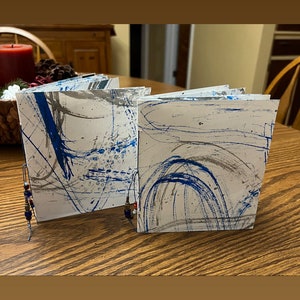 May include: Two small, handmade books with blue and gray abstract designs on the covers. The books are sitting on a wooden table and are held together by a beaded string.