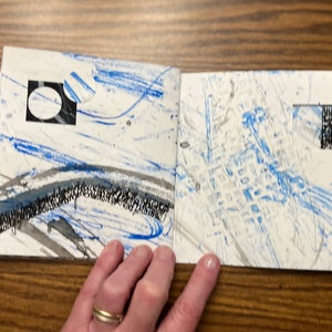 May include: An open book with blue and gray abstract art on the pages. The pages are white with blue and gray paint splatters and lines. There is a black square with a white circle inside on the left page.