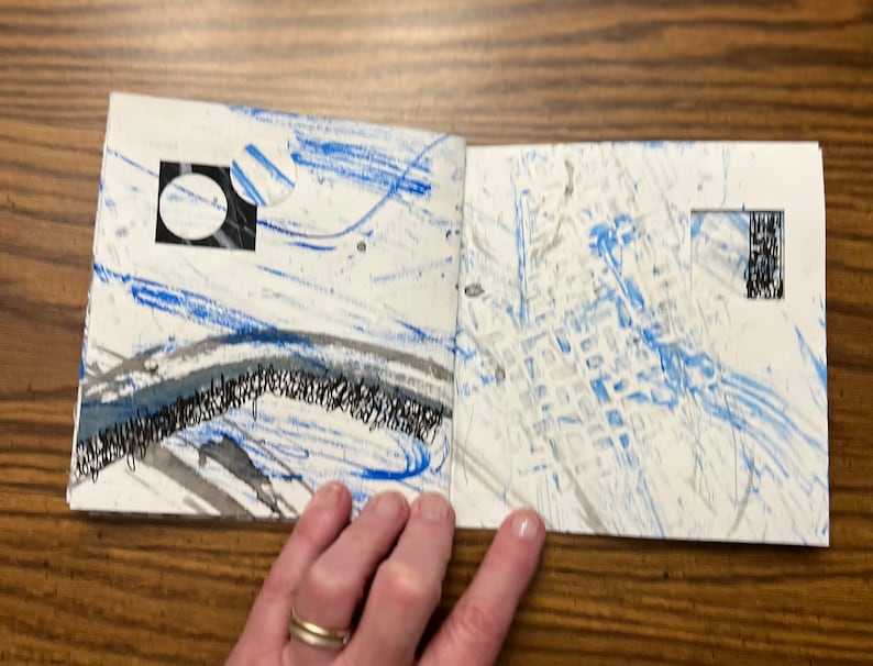 May include: An open book with blue and gray abstract art on the pages. The pages are white with blue and gray paint splatters and lines. There is a black square with a white circle inside on the left page.
