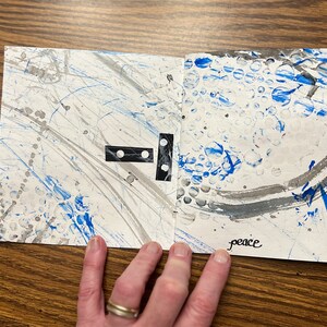 May include: A white paper journal with blue and gray paint splatters and the word "peace" written in black ink. The journal has a black and white domino piece on the left page.