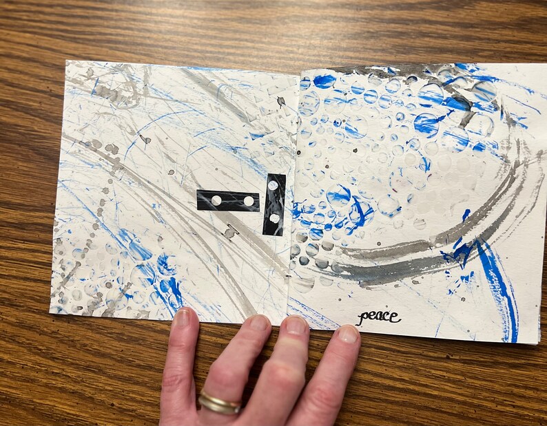 May include: A white paper journal with blue and gray paint splatters and the word "peace" written in black ink. The journal has a black and white domino piece on the left page.