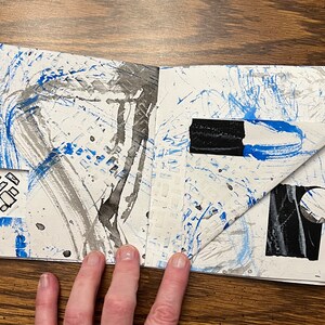 May include: An open sketchbook with blue and black paint splatters and brushstrokes on the pages. The pages are folded over to reveal a black and white circle and a black rectangle.