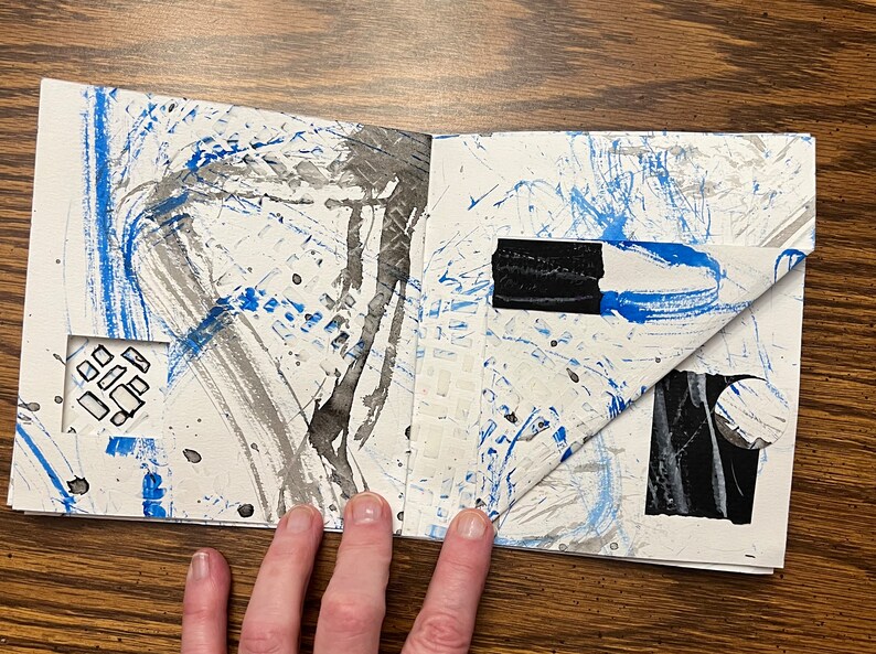 May include: An open sketchbook with blue and black paint splatters and brushstrokes on the pages. The pages are folded over to reveal a black and white circle and a black rectangle.