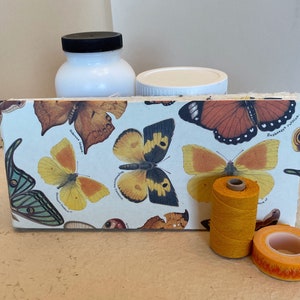 May include: A white journal with a butterfly design on the cover. The journal is surrounded by a spool of yellow thread and a roll of orange washi tape.