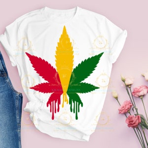 Puede incluir: Una camiseta blanca con un diseño de hoja de marihuana roja, amarilla y verde. La hoja está goteando pintura.