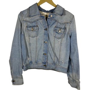 May include: Light blue denim jacket with a classic collar, button-down front, and two chest pockets. The jacket has a distressed look with subtle fading and wear marks. The sleeves have buttoned cuffs, and the jacket is hanging on a wooden hanger.