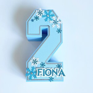 Puede incluir: Un adorno para pastel de n&uacute;mero dos azul con ribete plateado y decoraciones de copo de nieve. El nombre Fiona est&aacute; escrito en letras blancas en la parte inferior del adorno.
