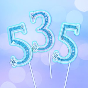 May include: Three blue and white cake toppers shaped like the numbers 5, 3, and 5. The numbers are decorated with glitter and snowflakes.