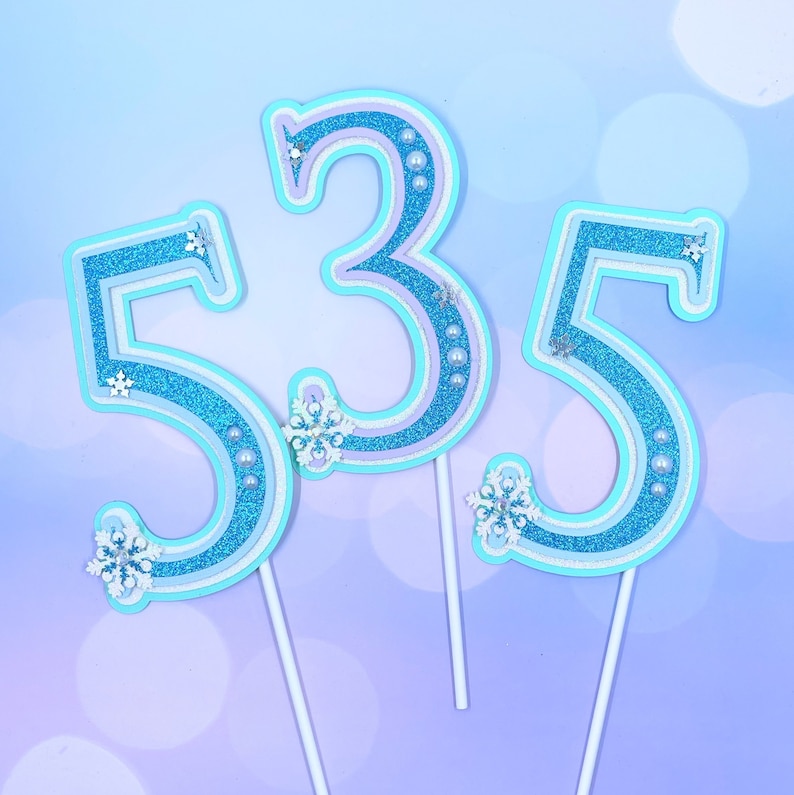 May include: Three blue and white cake toppers shaped like the numbers 5, 3, and 5. The numbers are decorated with glitter and snowflakes.