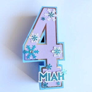 Puede incluir: Un adorno para pastel de n&uacute;mero cuatro de color morado claro con borde azul y detalles dorados. El n&uacute;mero est&aacute; decorado con copos de nieve blancos y azules y el nombre "Miah" est&aacute; escrito en letras azules con purpurina.