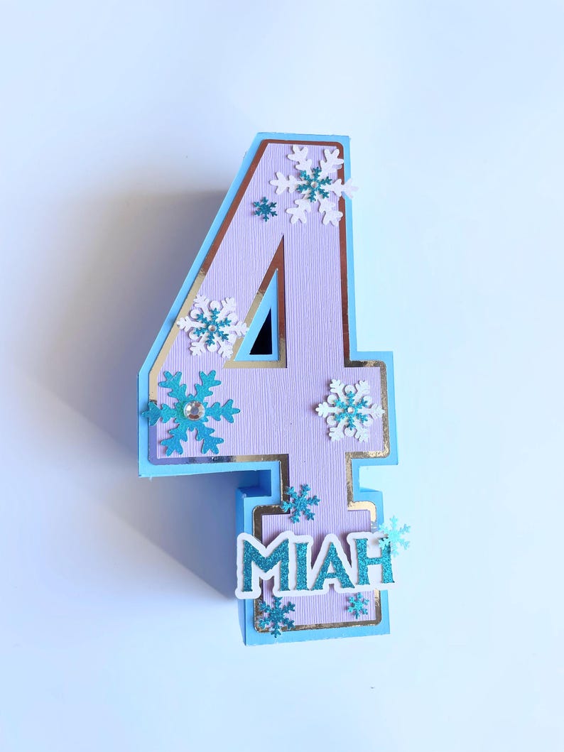 Puede incluir: Un adorno para pastel de n&uacute;mero cuatro de color morado claro con borde azul y detalles dorados. El n&uacute;mero est&aacute; decorado con copos de nieve blancos y azules y el nombre "Miah" est&aacute; escrito en letras azules con purpurina.