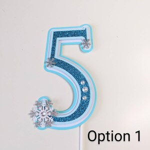 May include: A blue and white layered paper cake topper shaped like the number 5. The number is decorated with silver glitter and snowflake accents. The text "Option 1" is visible in the bottom right corner of the image.