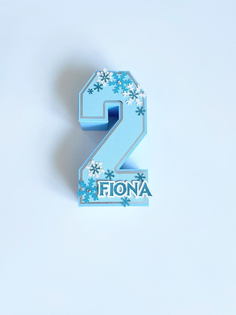 Puede incluir: Un adorno para pastel de n&uacute;mero dos azul con ribete plateado y decoraciones de copo de nieve. El nombre Fiona est&aacute; escrito en letras blancas en la parte inferior del adorno.