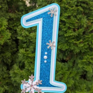 May include: A blue glitter number one cake topper with white accents and silver snowflake embellishments. The number one is layered and has a white outline.
