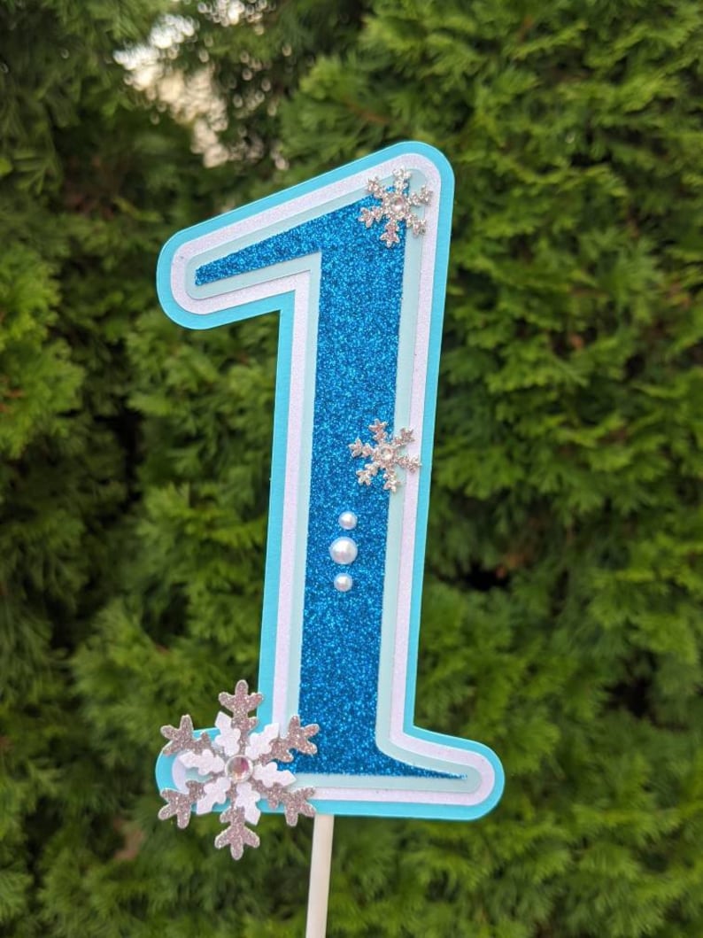 May include: A blue glitter number one cake topper with white accents and silver snowflake embellishments. The number one is layered and has a white outline.