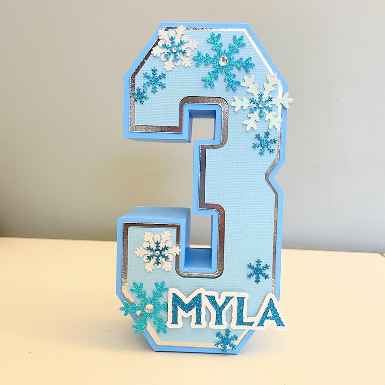 Puede incluir: Un n&uacute;mero tres en 3D azul y plateado con un borde blanco y un ribete plateado. El n&uacute;mero est&aacute; decorado con copos de nieve azules y blancos y el nombre "MYLA" est&aacute; escrito en letras blancas en la parte inferior.