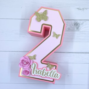 May include: A three-dimensional number two decoration in light pink with gold trim. It is adorned with four gold glitter butterflies and two layered pink paper flowers with pearl centers. The name Isabella is written in white script.