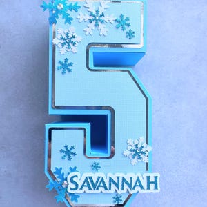 Puede incluir: Un n&uacute;mero cinco de papel azul y blanco con un borde plateado y decoraciones de copo de nieve. El n&uacute;mero est&aacute; decorado con el nombre "SAVANNAH" en letras blancas.