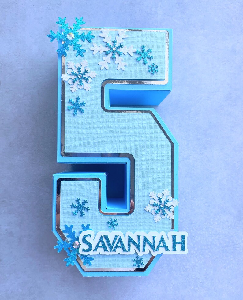 Puede incluir: Un n&uacute;mero cinco de papel azul y blanco con un borde plateado y decoraciones de copo de nieve. El n&uacute;mero est&aacute; decorado con el nombre "SAVANNAH" en letras blancas.