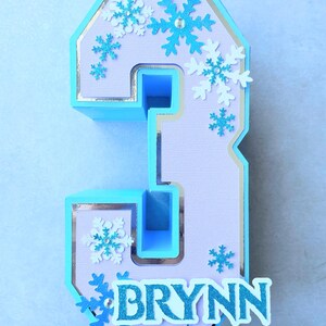 Puede incluir: Un n&uacute;mero tres azul claro y morado con un borde plateado. El n&uacute;mero est&aacute; decorado con copos de nieve blancos y azules y el nombre "BRYNN" est&aacute; escrito en letras azules con purpurina.