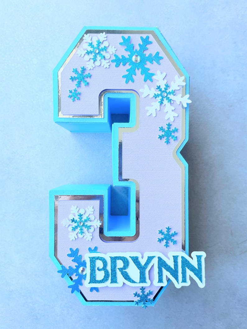 Puede incluir: Un n&uacute;mero tres azul claro y morado con un borde plateado. El n&uacute;mero est&aacute; decorado con copos de nieve blancos y azules y el nombre "BRYNN" est&aacute; escrito en letras azules con purpurina.