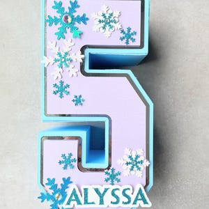 Puede incluir: Un n&uacute;mero cinco en 3D de color azul claro y morado con un borde plateado. El n&uacute;mero est&aacute; decorado con recortes de copos de nieve blancos y azules. El nombre "ALYSSA" est&aacute; escrito en letras blancas en la parte inferior del n&uacute;mero.
