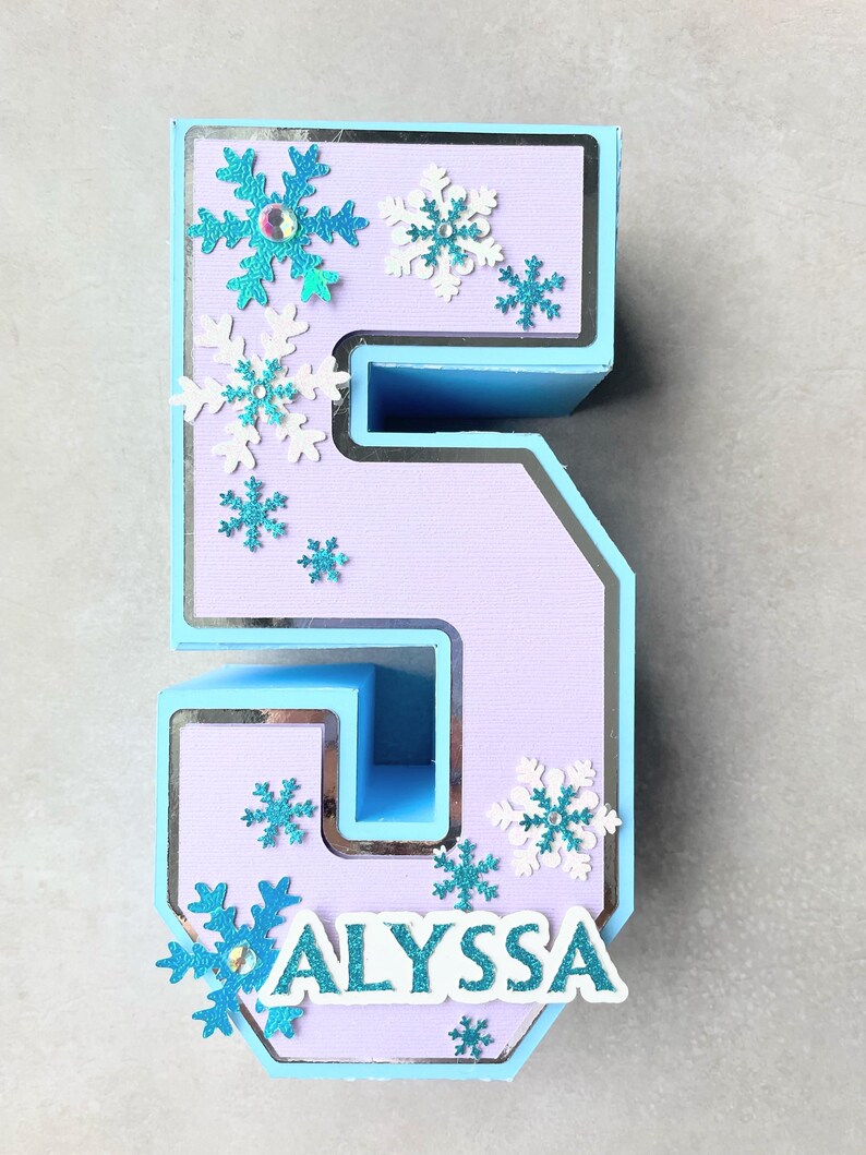 Puede incluir: Un n&uacute;mero cinco en 3D de color azul claro y morado con un borde plateado. El n&uacute;mero est&aacute; decorado con recortes de copos de nieve blancos y azules. El nombre "ALYSSA" est&aacute; escrito en letras blancas en la parte inferior del n&uacute;mero.
