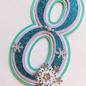 May include: A teal blue, purple, and white layered paper number eight with glitter and snowflake embellishments. The number is attached to a clear plastic stick.