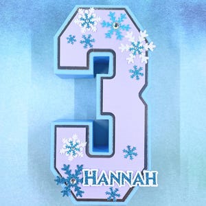 Puede incluir: Un n&uacute;mero tres azul claro y morado con un borde blanco. El n&uacute;mero est&aacute; decorado con copos de nieve azules y blancos y el nombre "HANNAH" est&aacute; escrito en la parte inferior.