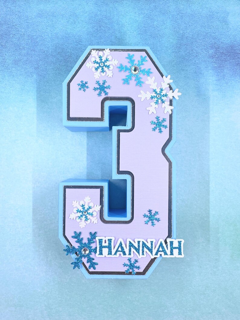 Puede incluir: Un n&uacute;mero tres azul claro y morado con un borde blanco. El n&uacute;mero est&aacute; decorado con copos de nieve azules y blancos y el nombre "HANNAH" est&aacute; escrito en la parte inferior.