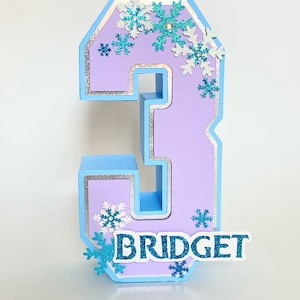Puede incluir: Un n&uacute;mero tres de color morado claro con un borde azul y purpurina plateada. El n&uacute;mero est&aacute; decorado con copos de nieve azules y blancos y el nombre "BRIDGET" est&aacute; escrito en purpurina azul en la parte inferior.
