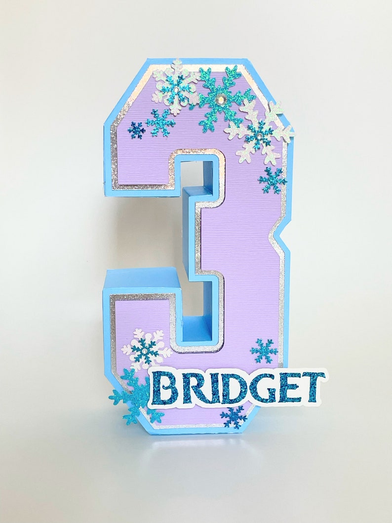 Puede incluir: Un n&uacute;mero tres de color morado claro con un borde azul y purpurina plateada. El n&uacute;mero est&aacute; decorado con copos de nieve azules y blancos y el nombre "BRIDGET" est&aacute; escrito en purpurina azul en la parte inferior.
