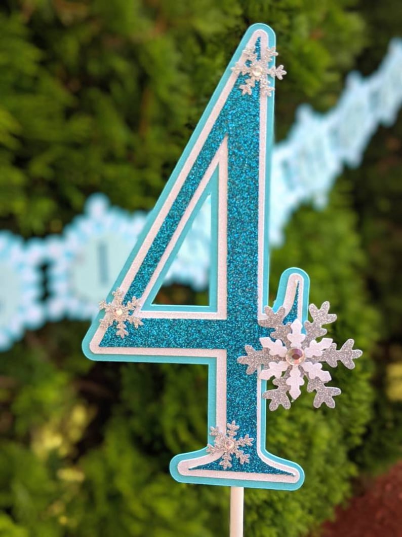 May include: A blue glitter number four with white outline and silver snowflake embellishments. The number is on a white stick.