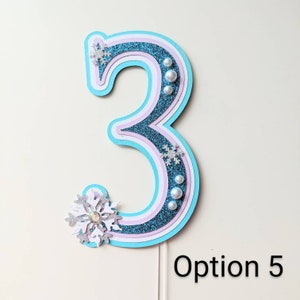 May include: A blue, pink, and white glitter number three cake topper with snowflake and pearl embellishments. Option 5.