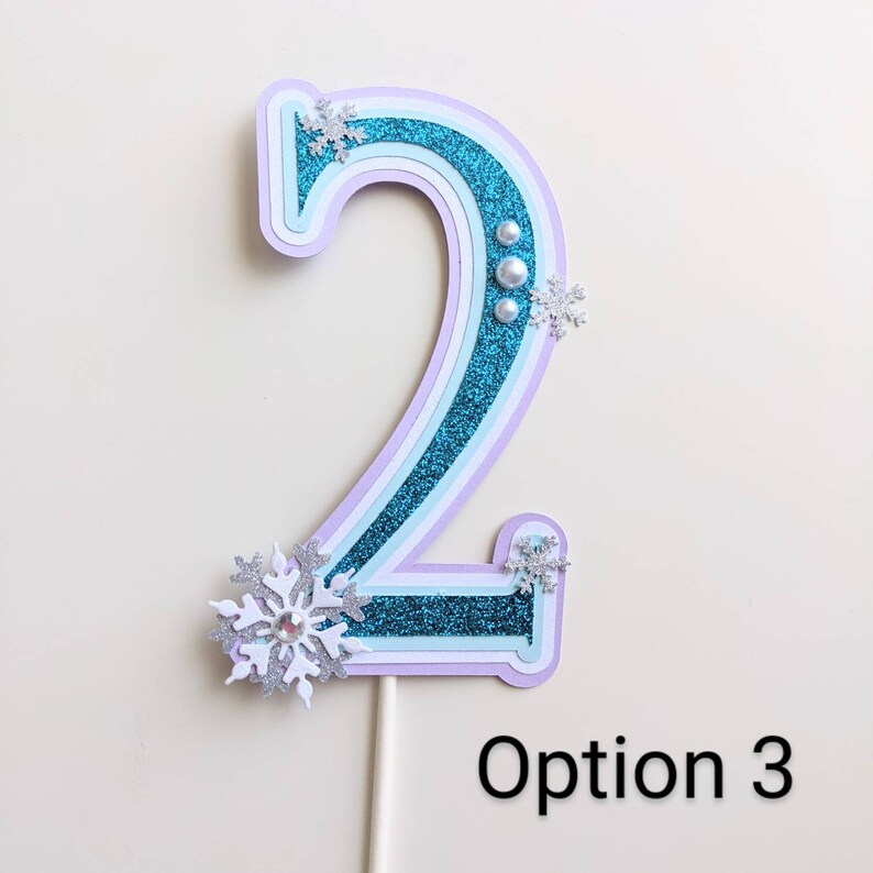 May include: A turquoise blue number two cake topper with white snowflake accents and silver glitter. The number is layered with white and purple paper and has three white pearl embellishments. The topper is on a white stick and the text "Option 3" is below the topper.