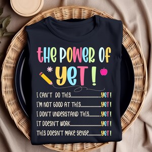 May include: A navy blue t-shirt with the text "the POWER of *YET!" in colorful letters. Below the text are phrases like "I CAN'T DO THIS...yet!" and "I'M NOT GOOD AT THIS...yet!" A pencil and an apple are also featured.