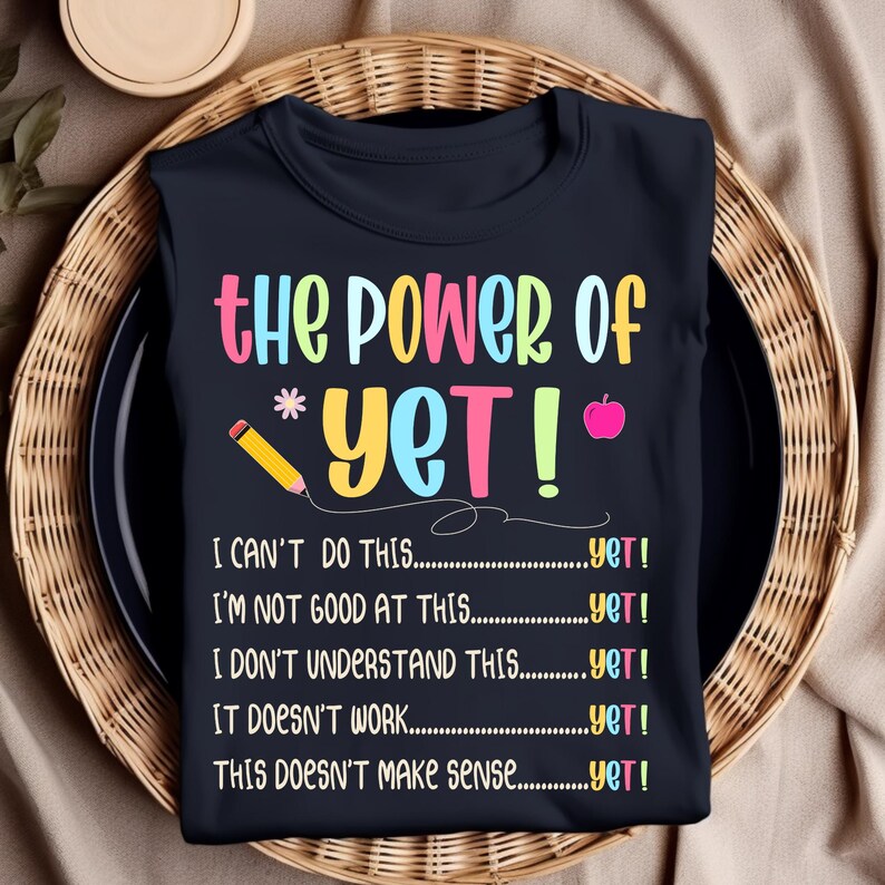 May include: A navy blue t-shirt with the text "the POWER of *YET!" in colorful letters. Below the text are phrases like "I CAN'T DO THIS...yet!" and "I'M NOT GOOD AT THIS...yet!" A pencil and an apple are also featured.