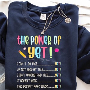 May include: Navy blue sweatshirt with the text "the Power of Yet!" in colorful letters. The design includes a pencil, an apple, and the phrases "I can't do this...yet!" and others. A silver necklace with a pendant is draped over the sweatshirt.