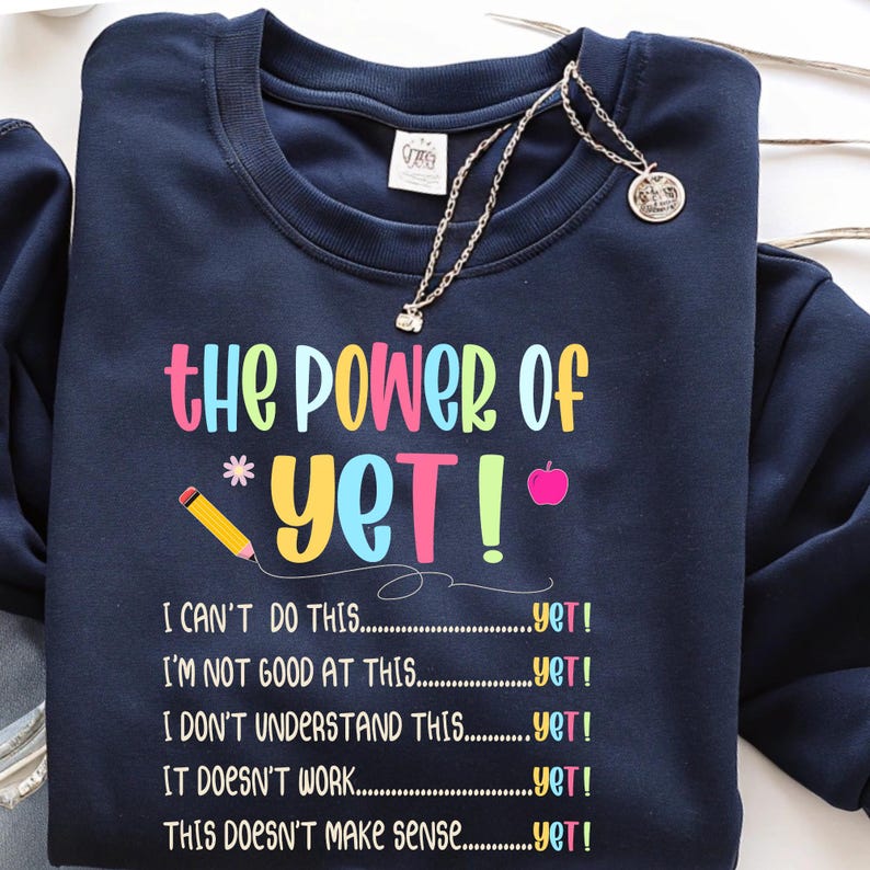 May include: Navy blue sweatshirt with the text "the Power of Yet!" in colorful letters. The design includes a pencil, an apple, and the phrases "I can't do this...yet!" and others. A silver necklace with a pendant is draped over the sweatshirt.