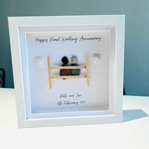 May include: A white framed picture with a pebble art design of a couple sitting on a bench. The text "Happy Pearl Wedding Anniversary" is at the top of the picture. The text "Pete and Jen 15th February 2023" is at the bottom of the picture.