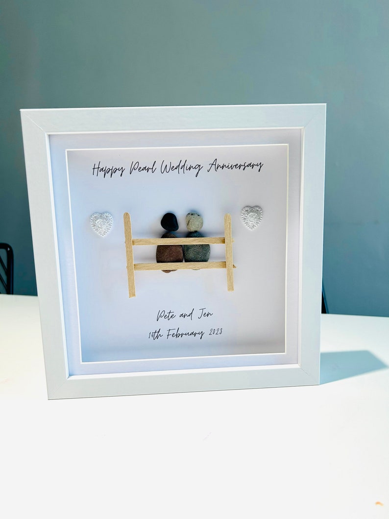 May include: A white framed picture with a pebble art design of a couple sitting on a bench. The text "Happy Pearl Wedding Anniversary" is at the top of the picture. The text "Pete and Jen 15th February 2023" is at the bottom of the picture.
