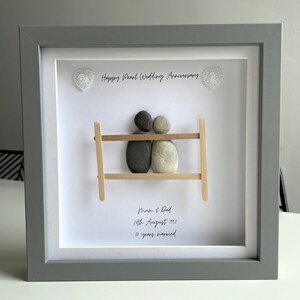 May include: A framed pebble art piece with two figures representing a couple sitting on a bench. The text reads "Happy Pearl Wedding Anniversary", "Mum & Dad", "28th August 1992", and "30 years married".