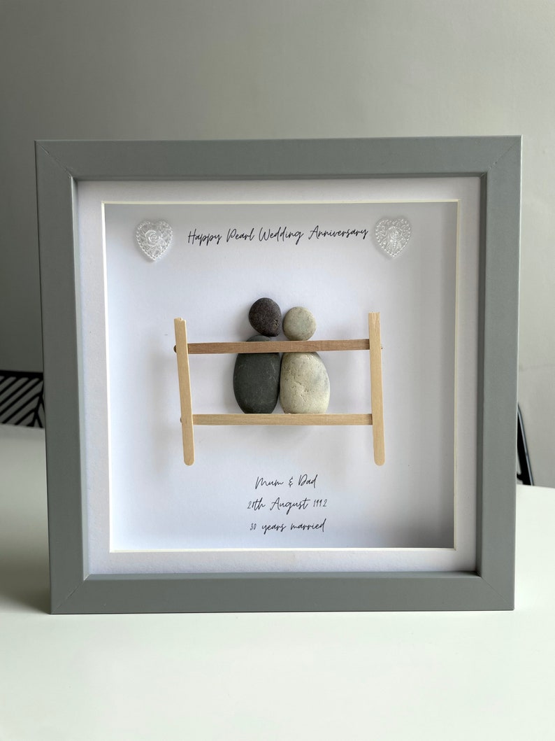 May include: A framed pebble art piece with two figures representing a couple sitting on a bench. The text reads "Happy Pearl Wedding Anniversary", "Mum & Dad", "28th August 1992", and "30 years married".