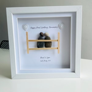 May include: A white framed picture with a pebble art design of a couple sitting on a wooden bench. The text "Happy Pearl Wedding Anniversary" is written above the couple, and "David & Lynn 2nd May 2022" is written below the couple.