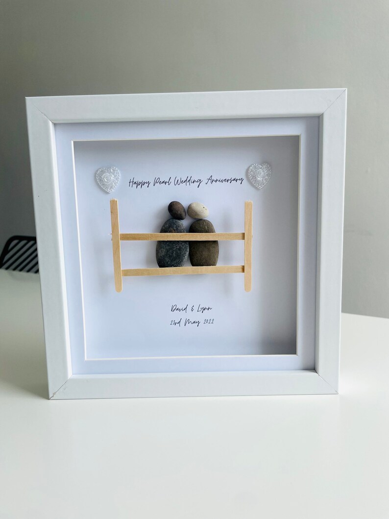 May include: A white framed picture with a pebble art design of a couple sitting on a wooden bench. The text "Happy Pearl Wedding Anniversary" is written above the couple, and "David & Lynn 2nd May 2022" is written below the couple.