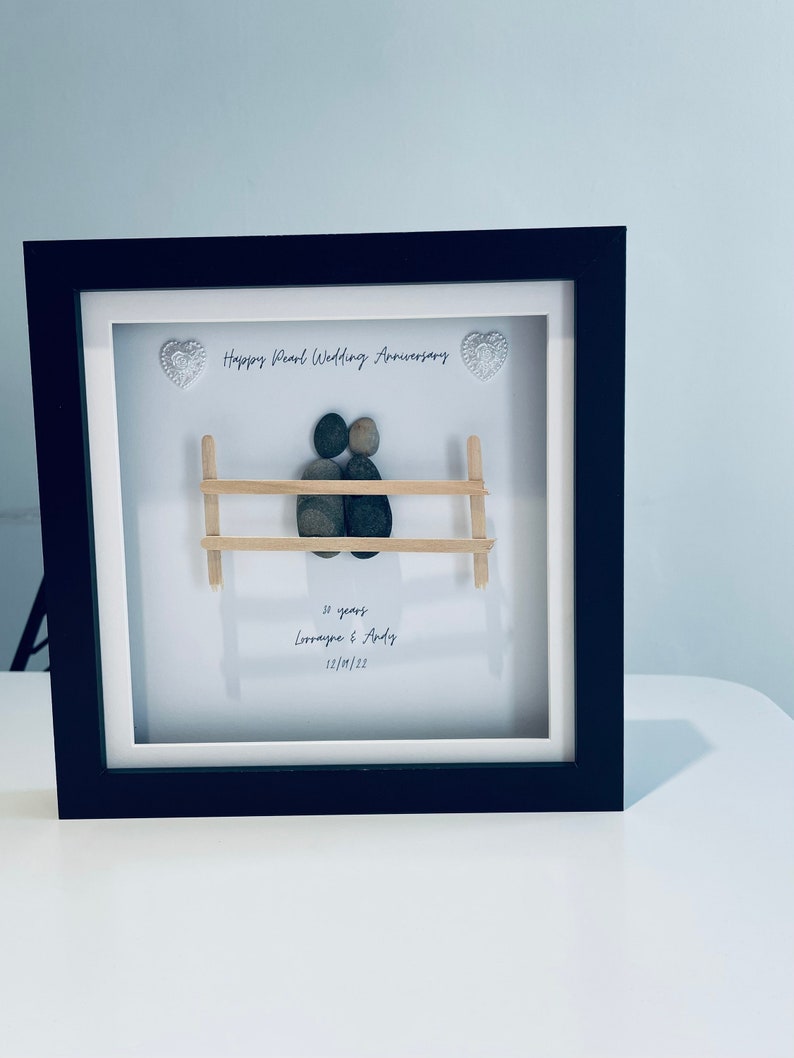 May include: A framed pebble art piece depicting a couple sitting on a bench. The art is titled "Happy Pearl Wedding Anniversary" and includes the text "30 years Lorraine & Andy 12/10/22".